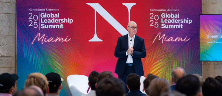 A man in a suit stands on stage addressing an audience at the 2025 Global Leadership Summit hosted by Northeastern University in Miami, with a colorful backdrop and large "N" behind him.
